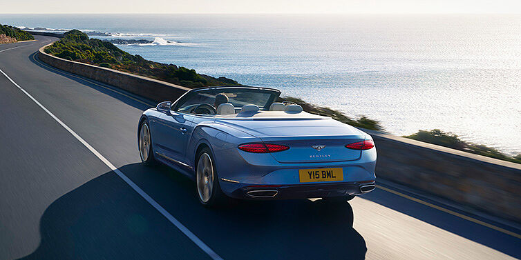 Bentley Hyderabad New Bentley Continental GTC Azure convertible rear view in Light Windsor blue paint with 22 inch ten spoke wheels driving dynamically on an ocean road in the sunshine